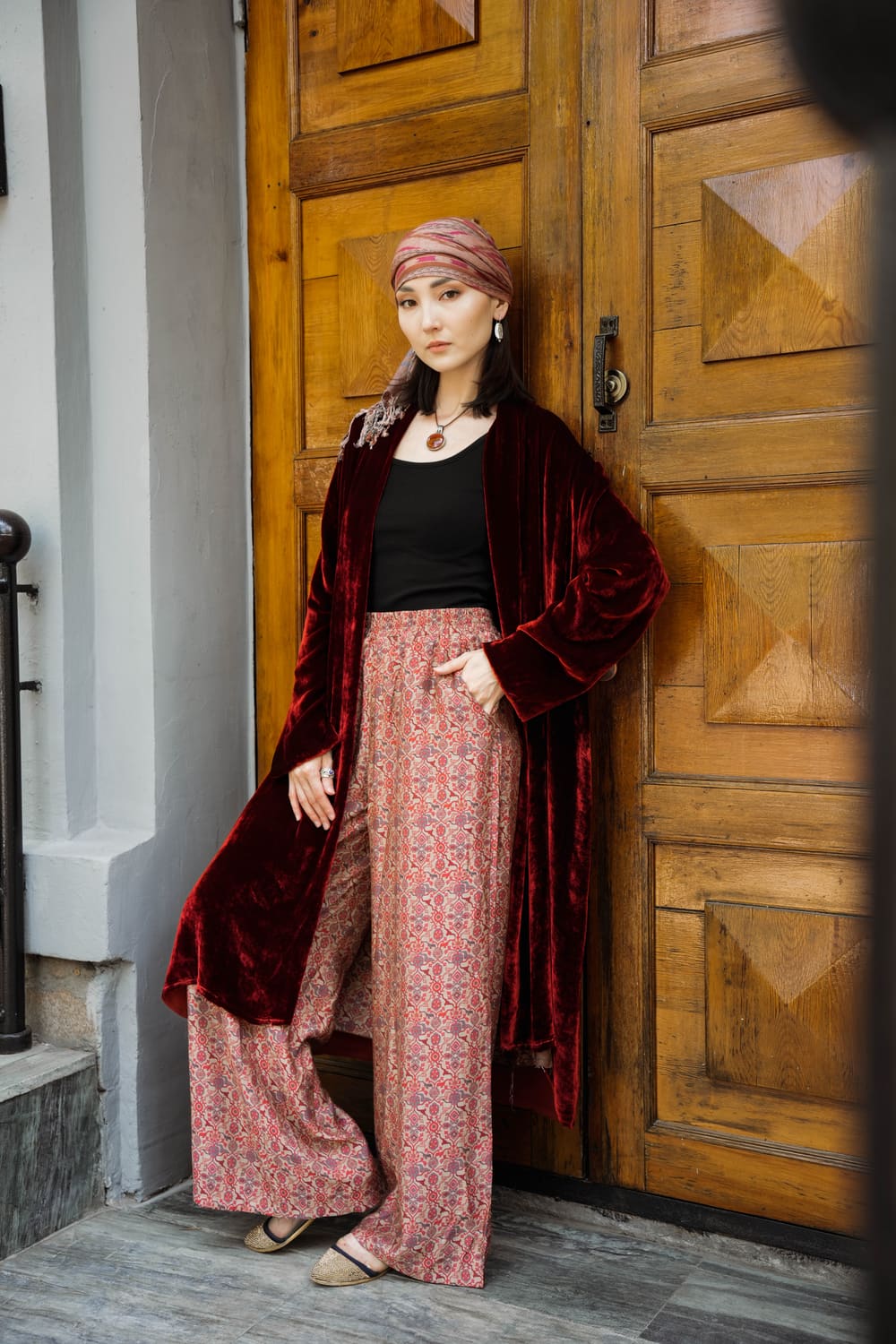 Burgundy velvet TURANAI chapan with finely printed trousers by a carved wooden door